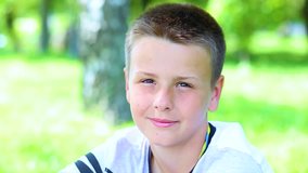 Closeup portrait of handsome caucasian young teenage boy isolated at green park background. Kid of 13 year old looks at camera smiling and winking  his eye cheerfully. Real time full hd video footage.