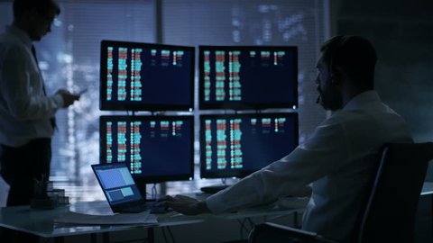 Late at Night in Trader's Bureau. Stockbroker Reads Numbers on His Multiple Displays with Stock Information on Them,He Also Consults Clients with Headset On.