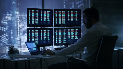Late at Night Trader Reads Numbers on His Multiple Displays with Stock Information on Them, He Also Consults Clients with Headset On. In Background Big City Window View.