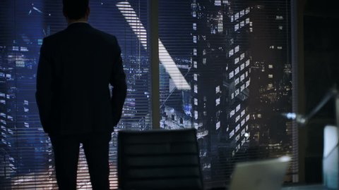 Late at Night Businessman Stands at the Window in His Office with Big City View. Then He Sits Down at His Desk and Starts Working on a Laptop. 