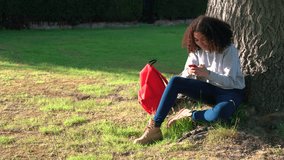 4K video clip of beautiful healthy mixed race African American girl teenager sitting by a tree with a red backpack using a cell phone sms text messaging 