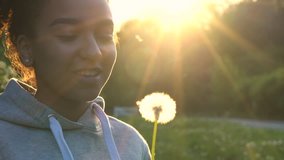 4K video clip of beautiful healthy mixed race African American girl teenager girl young woman laughing, smiling and blowing a dandelion at sunset or sunrise