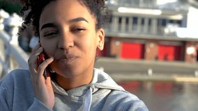 4K video clip of beautiful mixed race African American girl teenager young woman on a bridge over a river, talking on a mobile cell phone 
