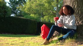 4K video clip of beautiful healthy mixed race African American girl teenager sitting by a tree with a red backpack using a cell phone sms text messaging 