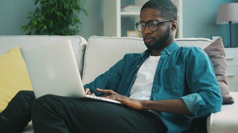 Attractive african American man in glasses sitting on the sofa using laptop at home living background for chatting with friends. Indoor. Technology,job, work Distant learning, lifestyle Online surfing