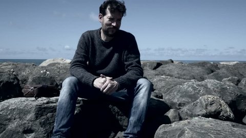 Sad man sitting on rocks in front of ocean slow motion desaturated