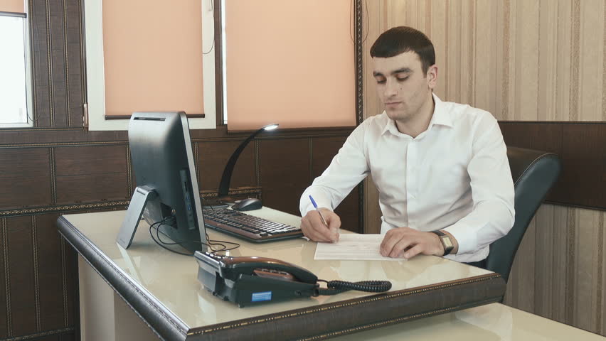 In a stylish office, a young man signs papers and works at a computer