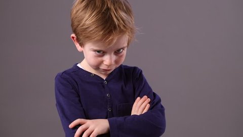 mischievous young 6-year old boy shaking his head and turning his back to refuse with a cheeky pouting smile and sulking body language, grey background indoor
