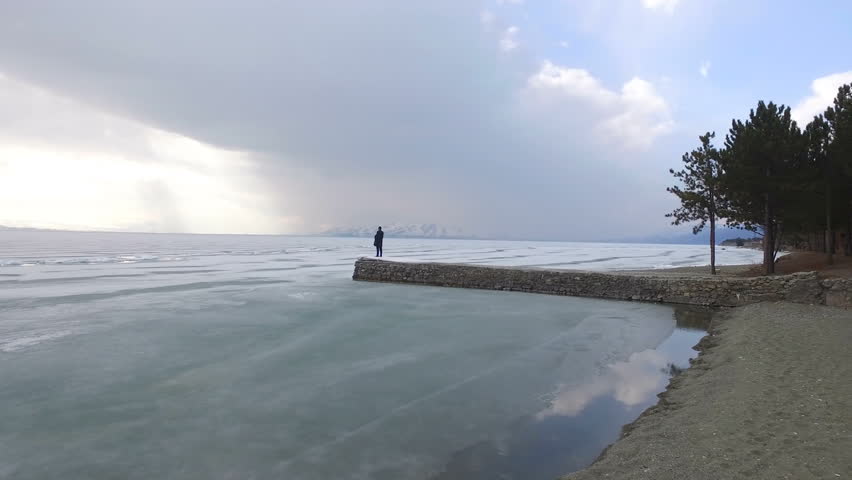 Aerial Flying near young man standing victoriously on pier to the iced lake. 