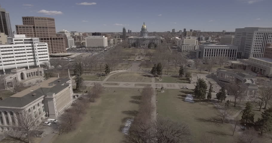Aerial of Downtown Denver Colorado fly through park to federal building. Snow covered mountains in background and city around. Captured Raw ready for color