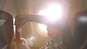 Closeup portrait of cute funny kid taking selfie in plane during flight. Child of 9 years sits on his seat near window with bright sunlight through it in background. Wide angle real time video footage