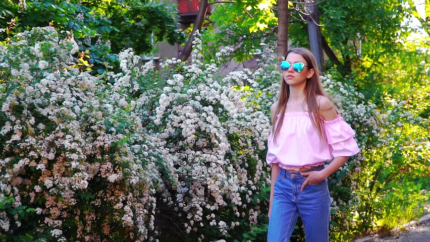 Teenager girl in pink shirt, blue jeans and sunglasses walking near the flowering bush at sunset and look up to the sky.