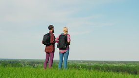 A man and a woman with backpacks standing at the edge of a large green hill or mountain. Looking forward to the horizon. Steadicam fly around video