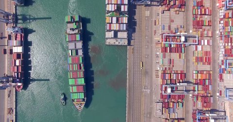 Commercial port with container ships during loading and unloading, and a container ship pulled by tugboats - Top down aerial view.