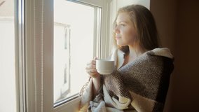 Closeup toned video of young woman covered in plaid drinking coffee at big window