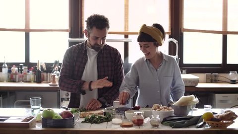 Funny happy couple cook dinner in an open space kitchen full of light, he is serious chops organic vegetables and she tries to steal a snack. they are excited, peaceful and loving