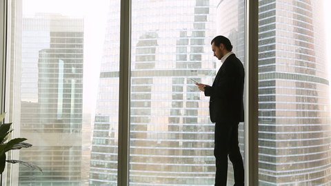 Confident young businessman wearing suit standing near full length window in office, holding tablet or smartphone, looking big city urban buildings, texting messages, chatting or using business apps