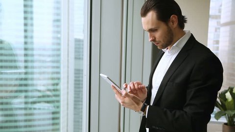 Attractive smiling businessman wearing suit holding using digital tablet, standing near office window, looking at skyscrapers, chatting texting messages, responding e-mails, working with business app