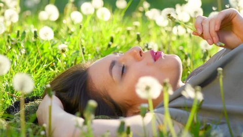 Beautiful Young Woman lying on the field in green grass and blowing dandelion. Outdoors. Enjoy Nature. Healthy Smiling Girl on spring lawn. Allergy free concept. 4K UHD video 3840X2160
