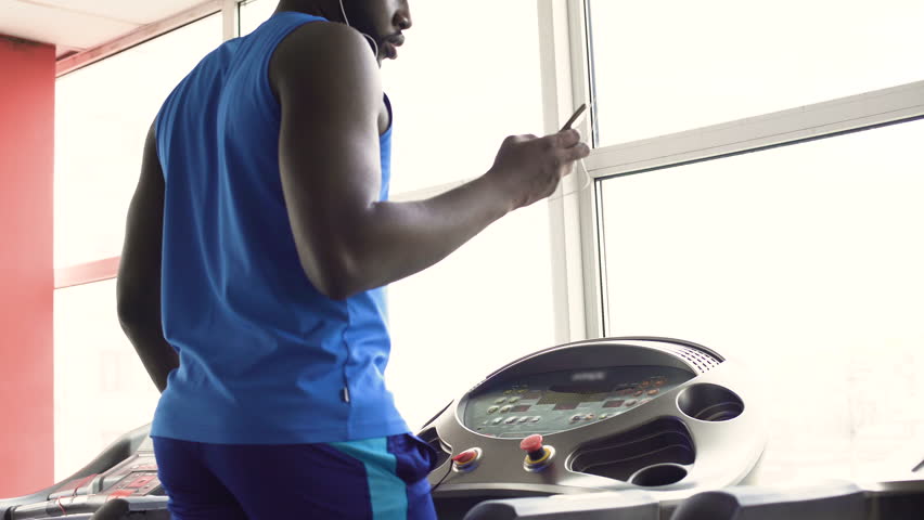 Afro-American male walking on treadmill and downloading fitness app on cellphone