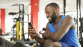 Happy African-American male sitting at the gym and watching video on cellphone