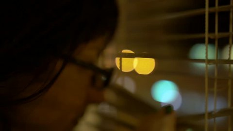 A young brunette at night with spectacles with a black rim comes to the window, pushes the blinds looks out the window carefully, gets frightened and leaves the window. Reflection in glasses, close-up