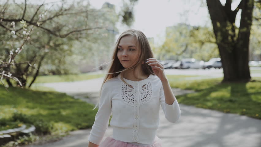 Pretty young woman in a pink skirt and wearing a white shirt wearing a smartphone in the park on a sunny day. Three-axis Gimbal stabilized tracking shot