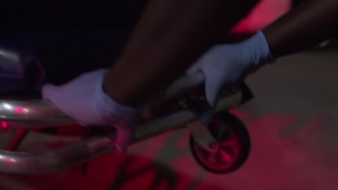Close-up tracking shot of coroner's gloved hands pushing a dead, bagged body on a gurney at a crime scene.