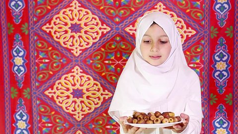 Little Muslim girl presenting a dish of dates for iftar - breaking fast in Holy Ramadan in front of islamic fabric background - showing generosity of Ramadan