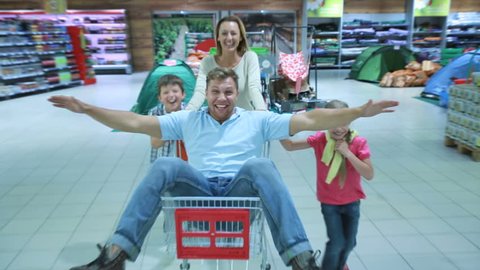 Mom and two kids having fun wheeling daddy around store in the market shopping cart., videoclip de stoc