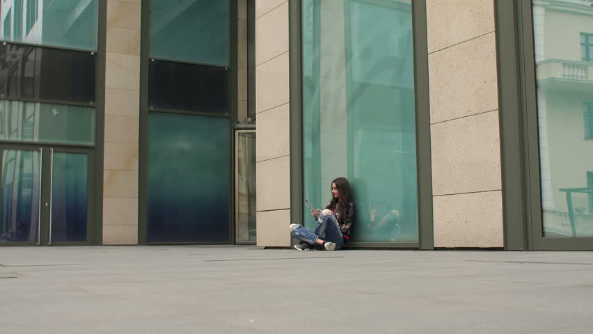 Fashionable girl in ripped jeans sitting on the ground with the tablet leaning on a modern glass building.