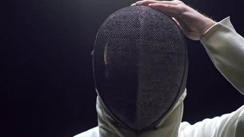 Tilt up shot of young man in white costume holding fencing foil and putting protective mask in slow motion
