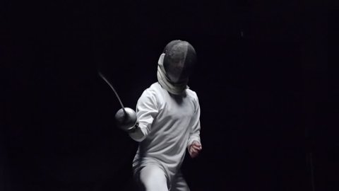 Slow motion shot of professional fencer in protective uniform practicing maneuvers with foil alone in dark studio