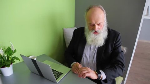 Elderly Smart Man Views and Learns New Technologies, Checks Capabilities of Iwatch and Understands Characteristics, Sitting at Office Table in Office of Company During Day. Man With Long Gray Beard
