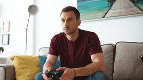 Attractive young man in T-shirt, playing console and sits on the couch at home, feeling happy