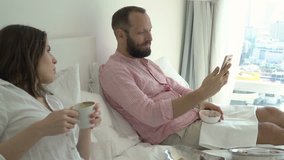 Young couple with smartphone talking during breakfast on bed at apartment
