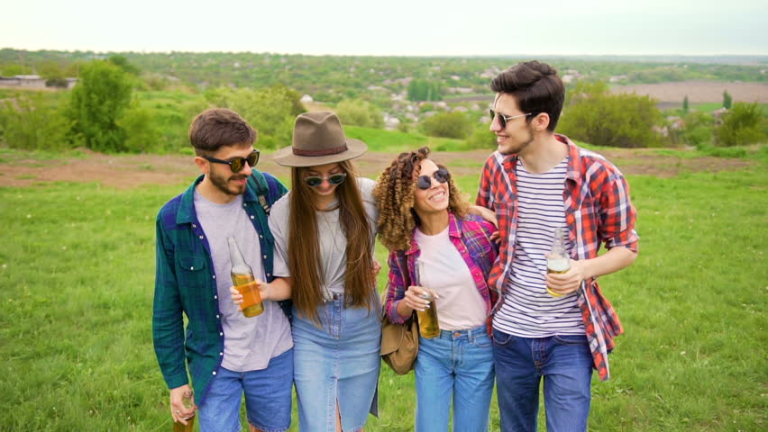 Four stylish happy hipsters are walking in the park with bottles of beer, embracing together. They clink with bottles. Slow motion medium shot. Royalty-Free Stock Footage #27133936