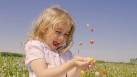 Cute little girl with pleasure catches multicolored candy falling from above. Joyful cheerful child laughing outdoors. Summer sunny day. Slow Motion.