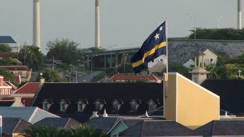 View from a ship as the camera tracks the flag of Curacao. Large industrial chimneys are in the background