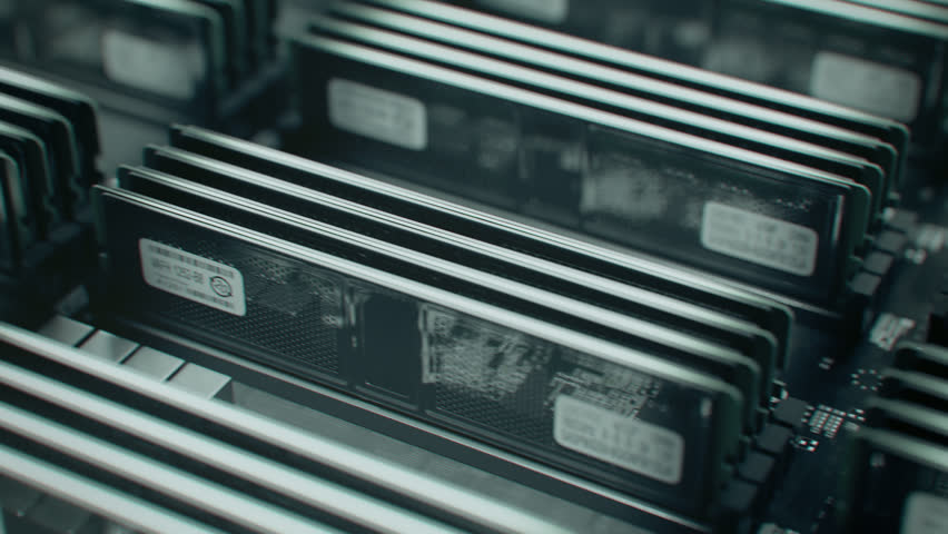 A slow close-up shot moves across rows of black storage drives mounted in a server chassis, with metallic heat sinks and barcode labels under dim industrial lighting. The shallow depth of field keeps the central drives sharp while the surrounding rack blurs into the background.