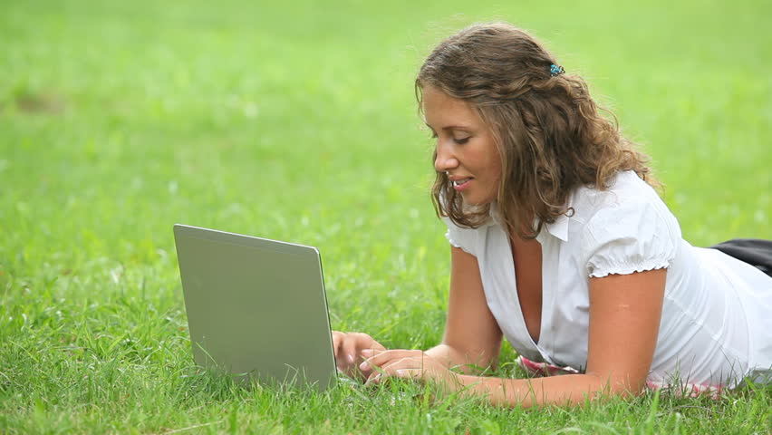 Young pretty businesswoman lying on green grass and typing on laptop