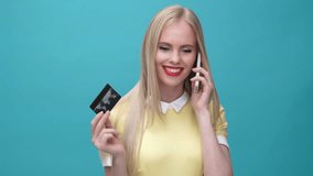 Young smiling woman with credit card talking on phone about purchases and winking isolated