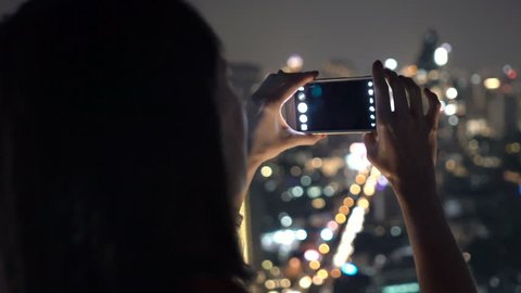 Woman taking photo of cityscape view with cellphone in bar at night 
