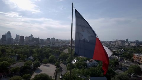 Large Texas flag waving while camera orbits with downtown Austin and the state capital building in the background. Aerial perspective.