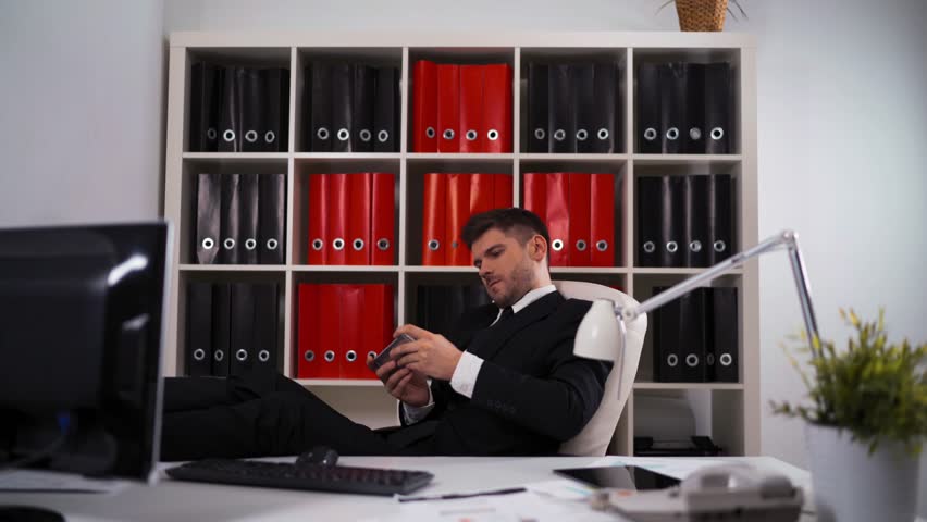 Ultra HD Young attractive businessman playing in game on the mobile smart phone in modern cool office on armchair with lamp computer digital pad keyboard. teal and orange.