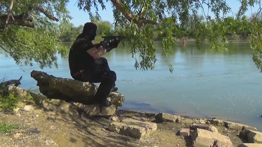 A Russian special forces soldier in a balaclava, sitting on a rock near the shore of the lake and aiming from a crossbow. Marine recon.