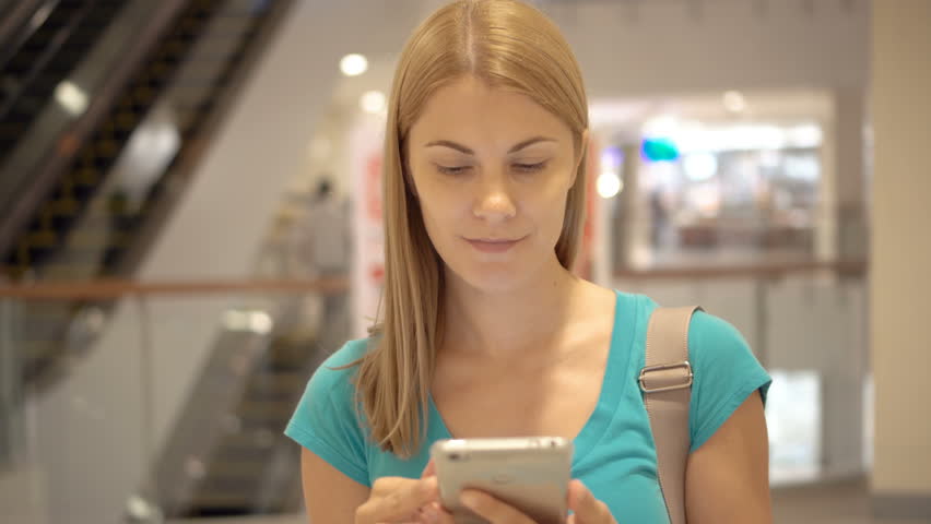 Young smiling woman in blue t-shirt standing in shopping center using smartphone, browsing, reading news, chatting with friends