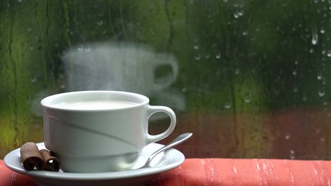 Cup with steaming hot milk is in front of the window. Winter and rain are outside.