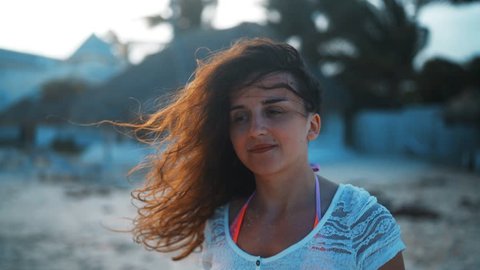 Close up portrait of young seductive brunette woman smiling on camera and laugh, hair blowing in wind. Sunny beach background at sunset. 4k ultra hd handy shot.