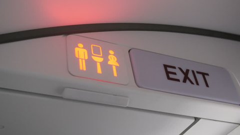 Shot of airplane lavatory sign turning from red (occupied) to green (available). Beside the lavatory sign is an exit sign.